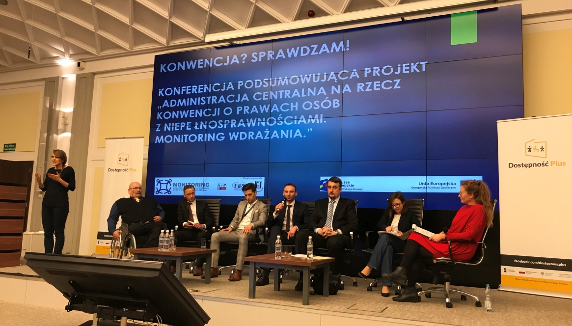 Sześcioro uczestników i prowadząca panel członkini jednego z zespołów monitorujących siedzą na podium. W środku z mikrofonem w ręku dyrektor Marcin Cudak. Z boku stoi tłumacz migowy wykonujący tłumaczenie.