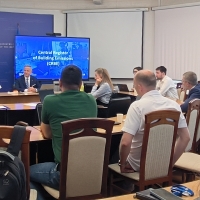 Tomasz Trojan, naczelnik, podczas prezentacji w imieniu Departamentu Usług Cyfrowych. Siedzi przy biurku, przed którym stoją flagi Mołdawii i Polski. Za nim znajduje się niebieskie tło z napisem 'MINISTERUL ENERGIEI REPUBLICII MOLDOVA' oraz herbem Ministerstwa Energii Mołdawii. Po jego prawej stronie widoczny jest duży ekran z wyświetlonym systemem poczty elektronicznej lub zarządzania dokumentami. 