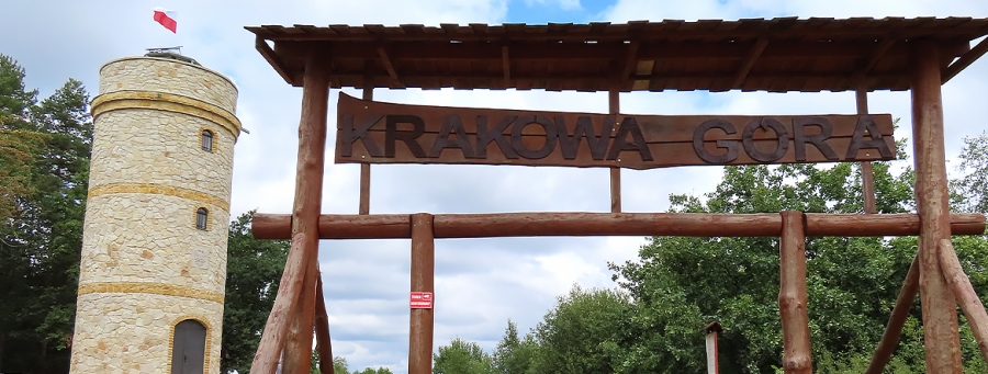Na zdjęciu widać kamienną wieżę z biało-czerwoną flagą na szczycie. Na pierwszym planie stoi drewniana brama z napisem Krakowa Góra.