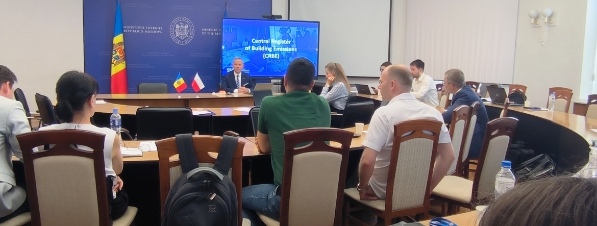 Sala konferencyjna z uczestnikami siedzącymi wokół dużego, zakrzywionego stołu. Na przodzie mężczyzna przemawia z mównicy, za którą znajdują się dwie flagi: Mołdawii i Polski. Na ekranie za nim widnieje napis 'Central Register of Building Emissions (CRBE)'. Pomieszczenie jest jasne, z białymi ścianami i naturalnym światłem wpadającym przez okna po prawej stronie.